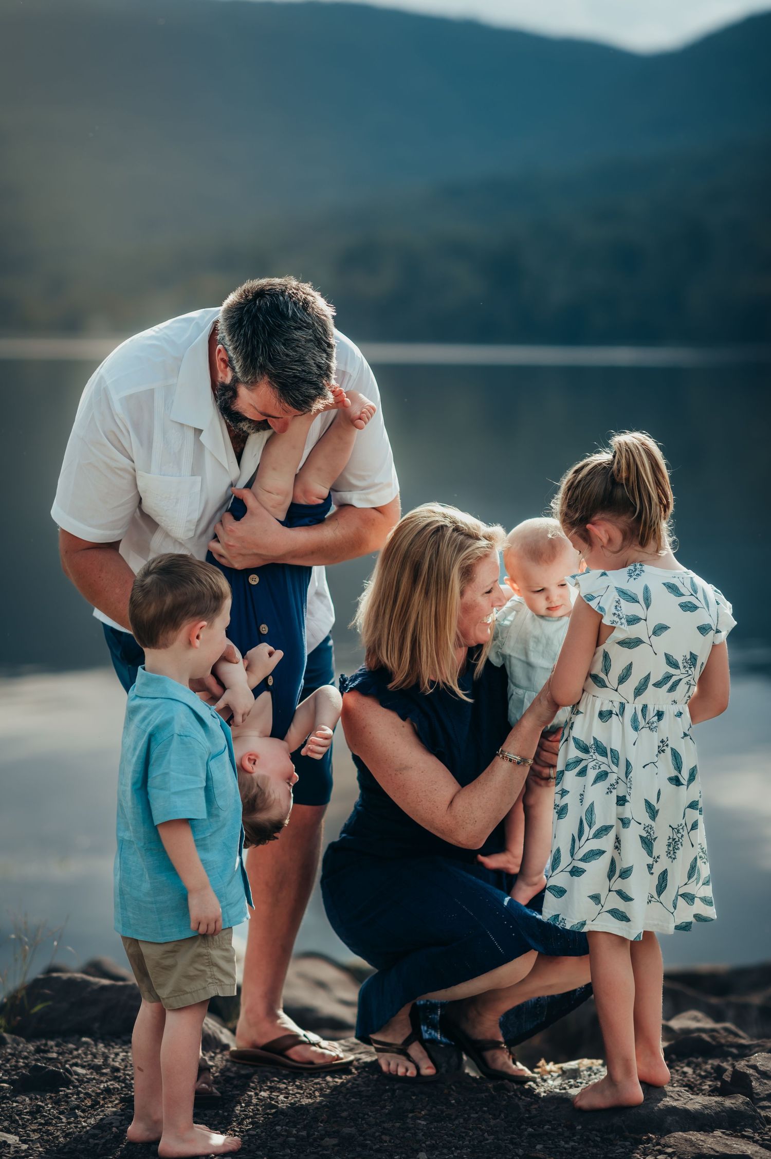 Dad holds baby boy upside downn for big brother to tickle while mom interacts with 2 daughters