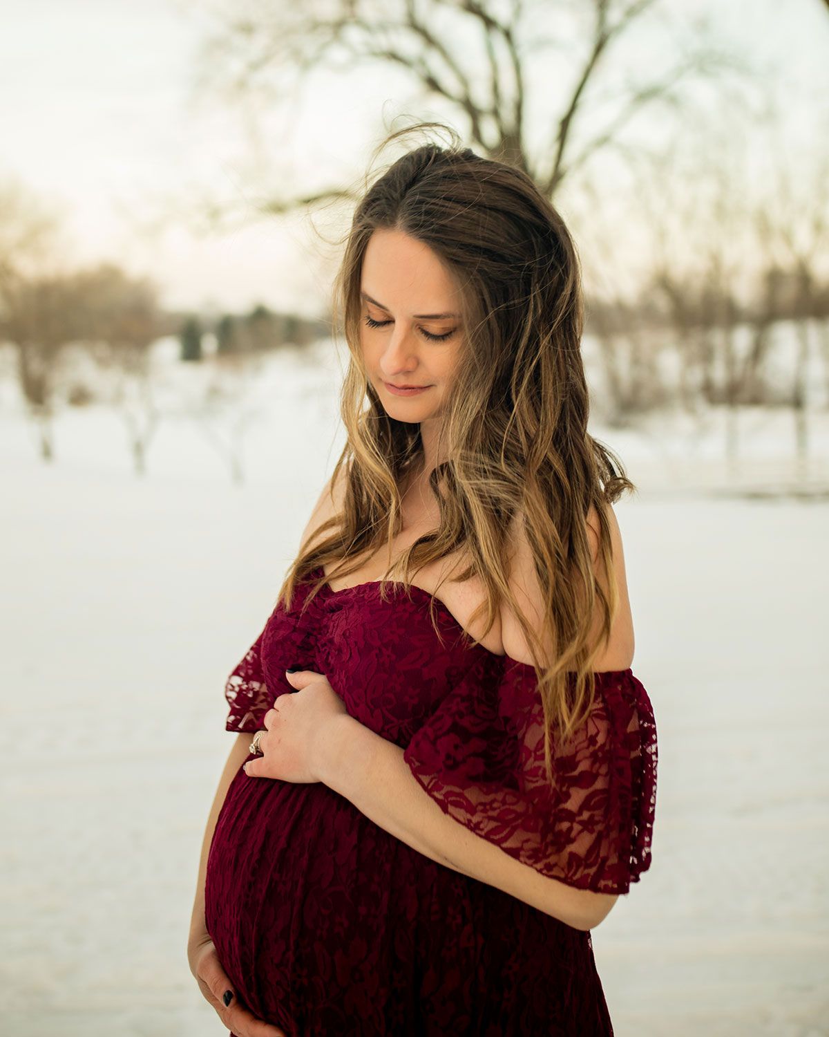 Woman in off-shoulder burgundy dress stands in snowy setting during portrait session.