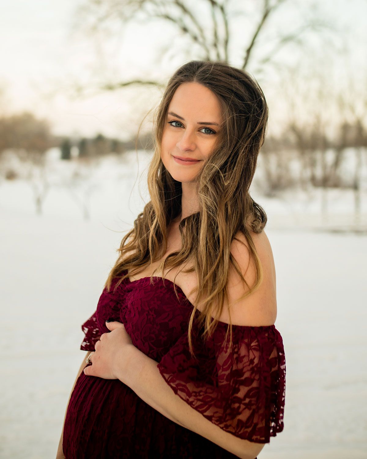 Woman in burgundy lace dress poses against snowy winter landscape.