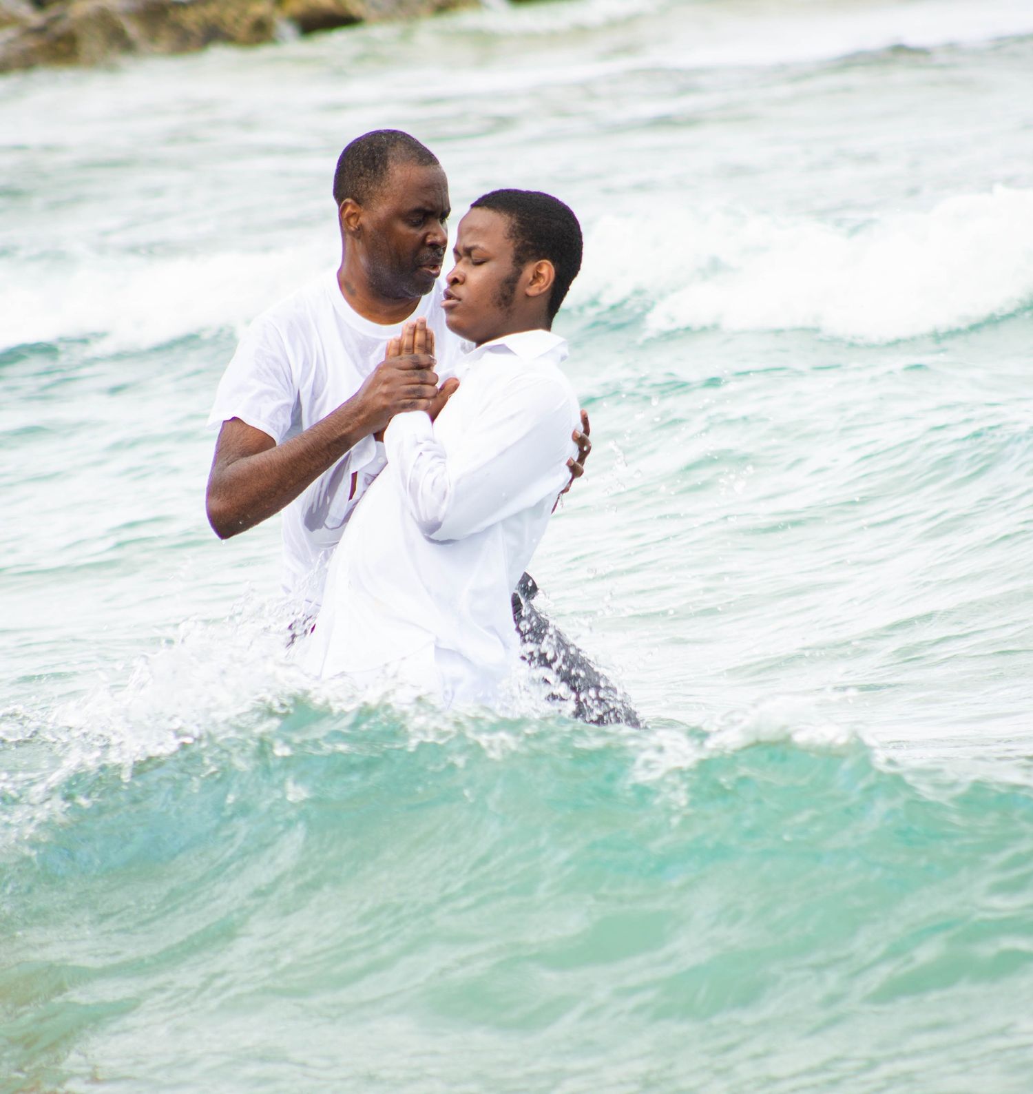 Two people embracing while standing in turquoise ocean waves wearing white clothing.