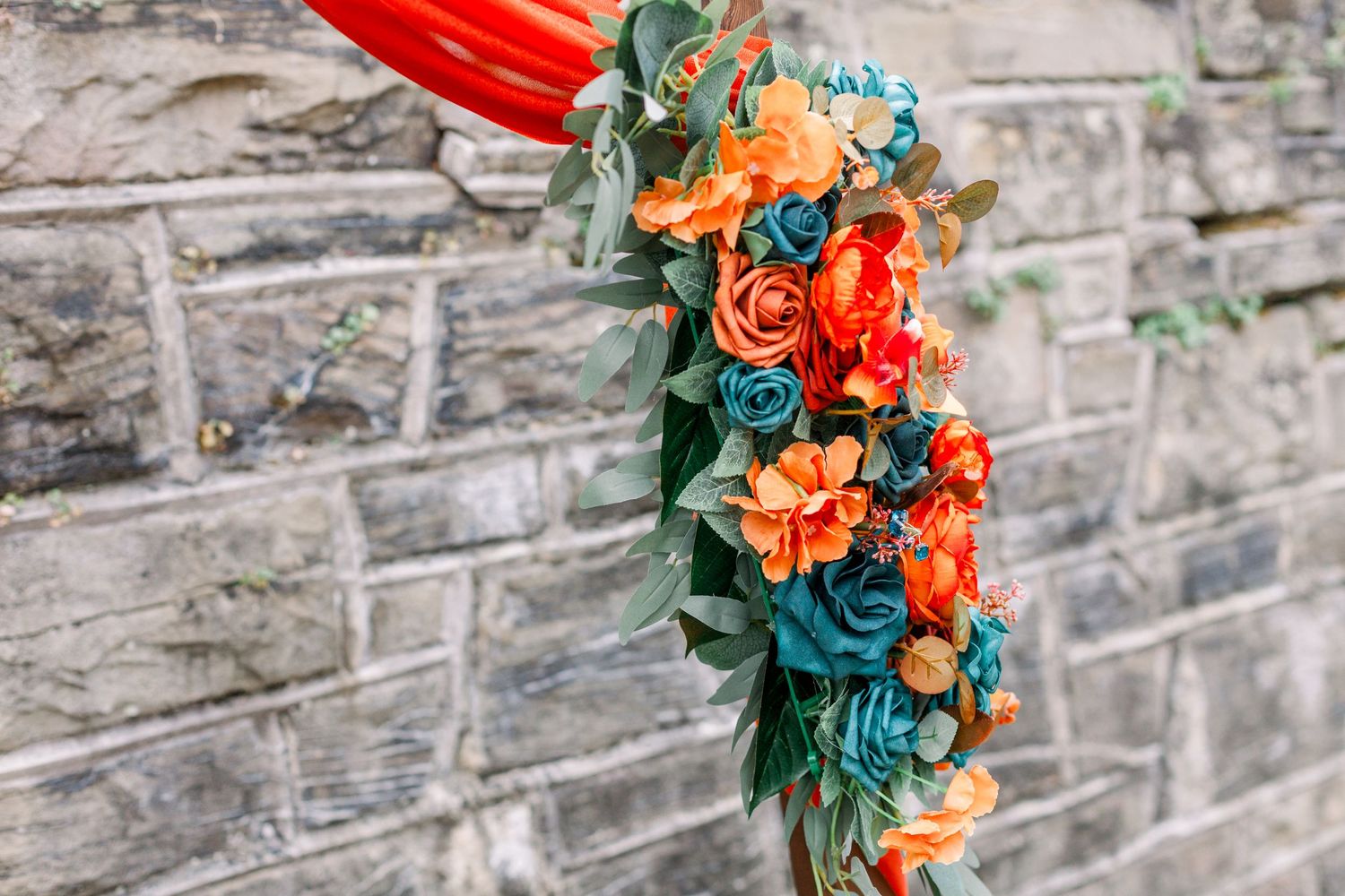 Colorful floral arrangement with orange and teal blooms against brick wall.