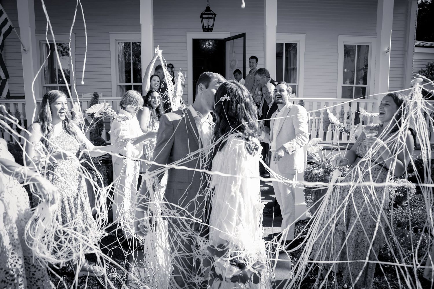 Black and white photograph of wedding guests gathered on a porch with tall grass in foreground.