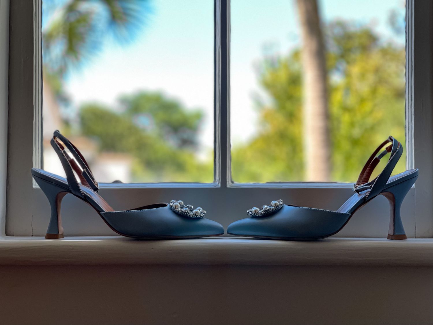 Blue wedding shoes with decorative details displayed on a windowsill with palm trees visible outside.