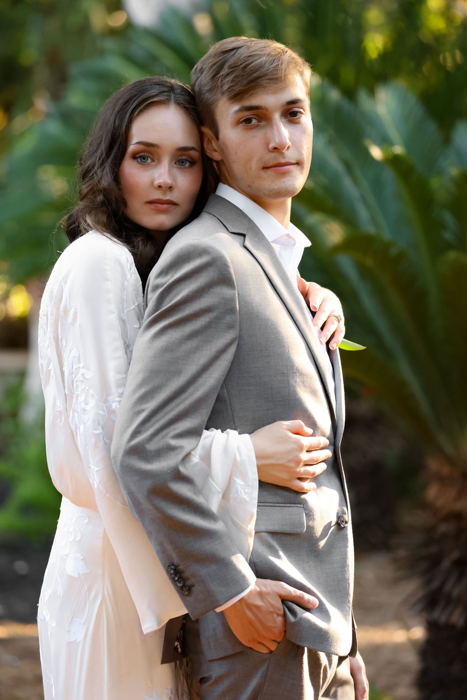 A romantic portrait of a couple in white and grey formal attire against a lush garden background.