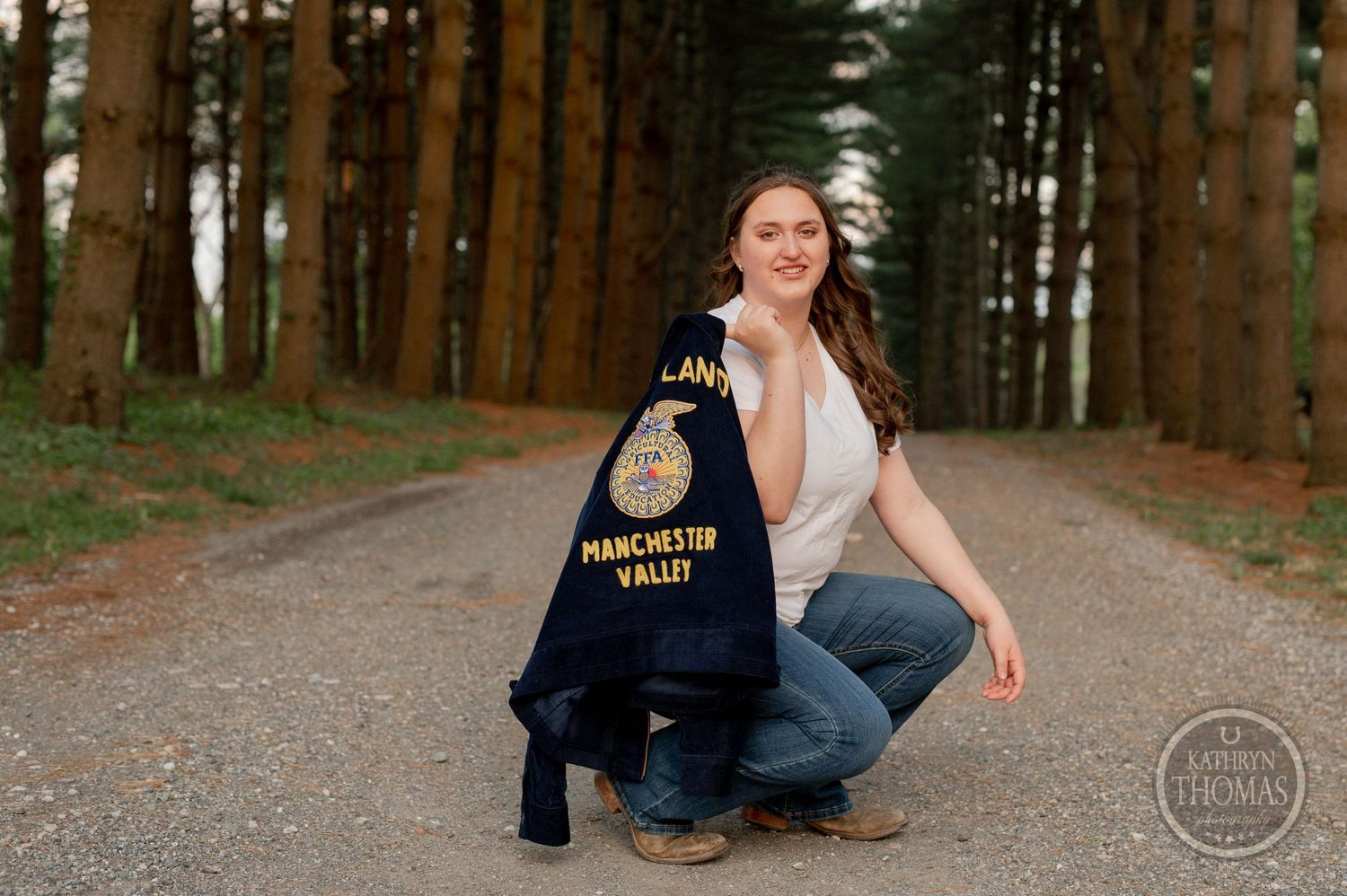 Person in jeans crouches on a pine-lined path displaying an FFA jacket.
