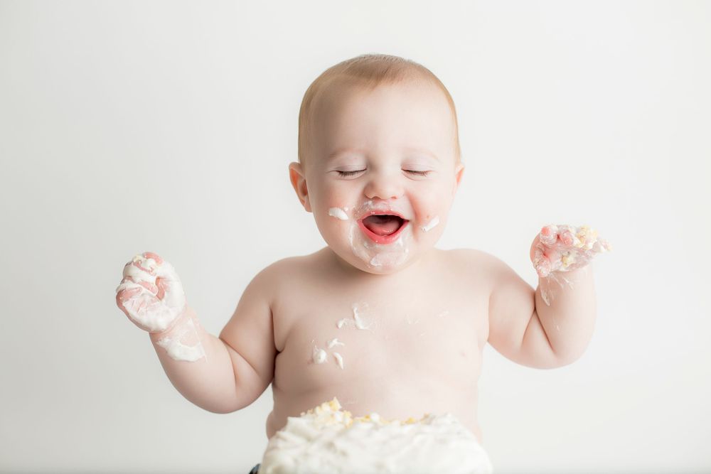 One year old baby boy with red hair laughing with cake on hands and mouth.