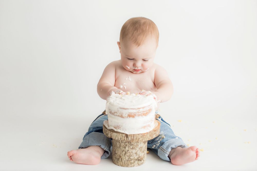 One year old baby boy sitting on a white backdrop wearing jeans and suspenders with cake on a tree cake stand.