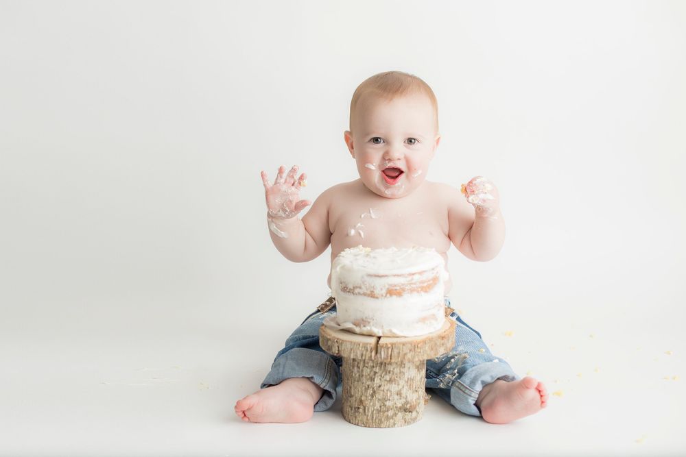 One year old baby boy smiling, sitting on white backdrop with cake in-between his legs.