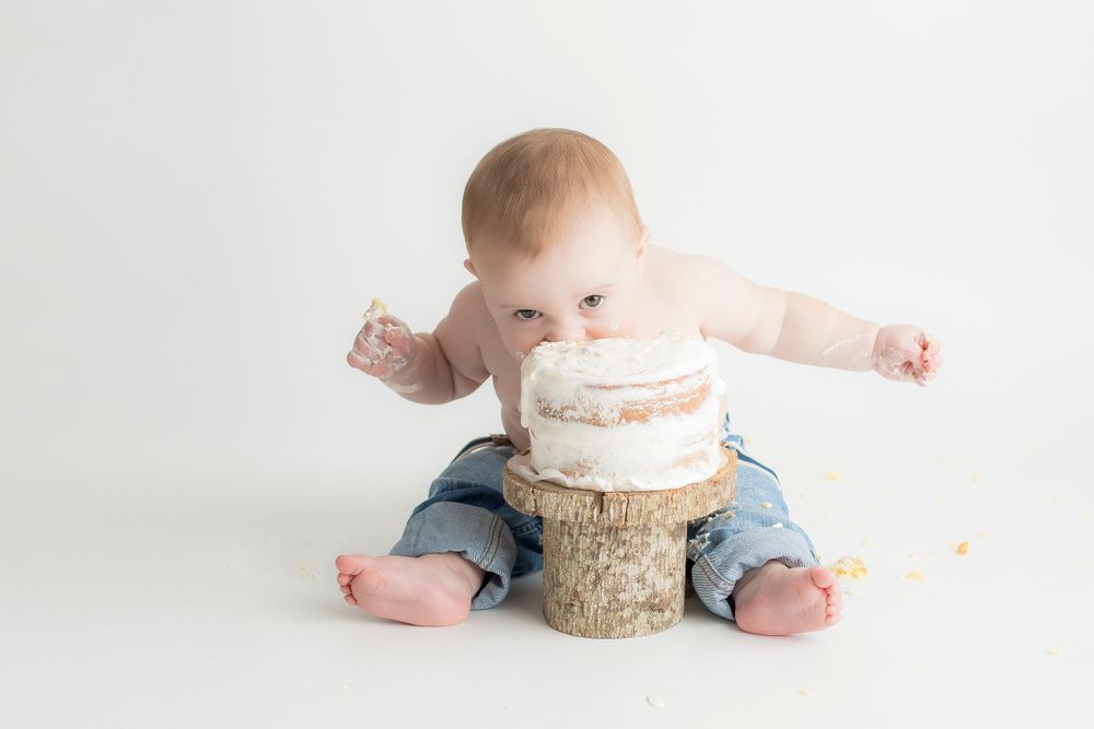 One year old baby boy sitting on white backdrop with cake in front of him.  Eating the cake with his mouth.