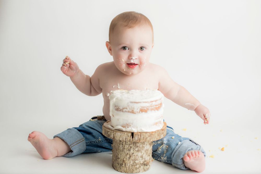 One year old baby boy sitting with cake between his legs.  Icing is on his nose.