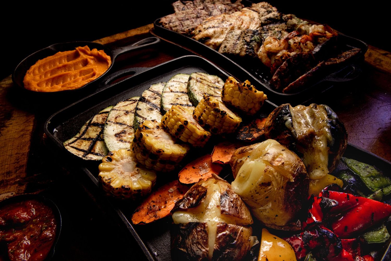 Grilled meats and vegetables arranged on dark platters with dramatic lighting.