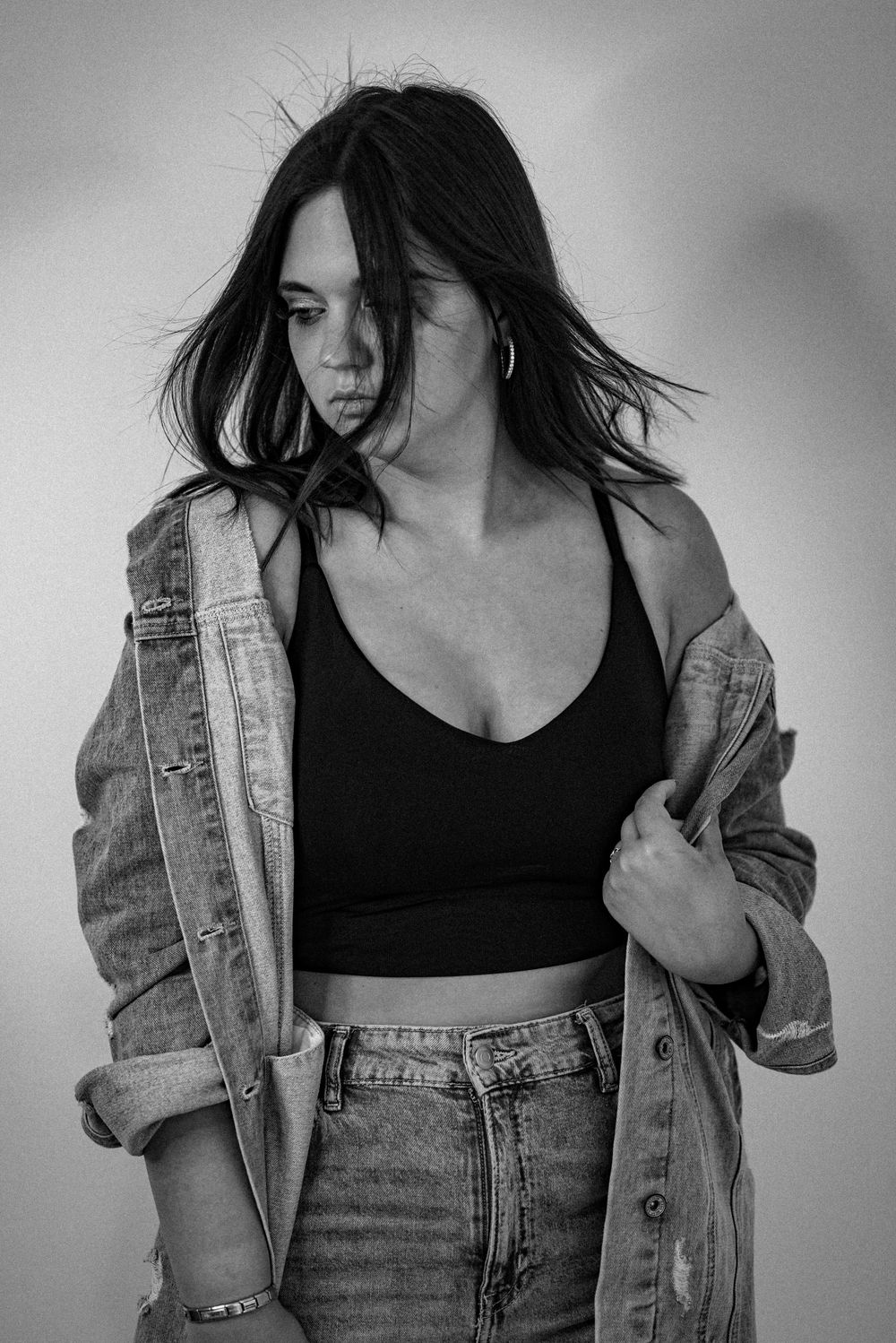 Artistic black and white portrait of a model in denim jacket and crop top with windswept hair against blurred background.
