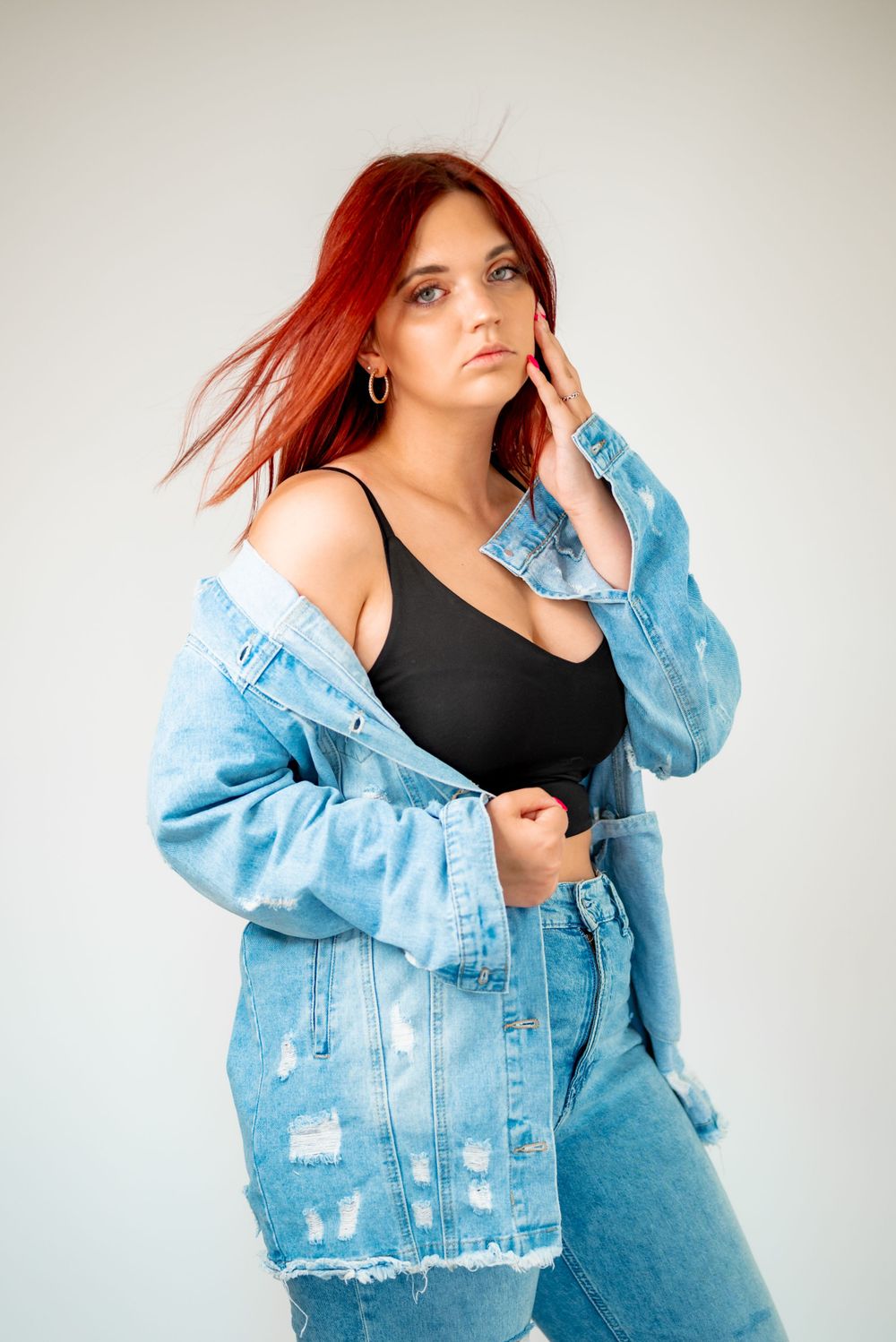 Fashion model in black top and light denim jacket posing against white backdrop.