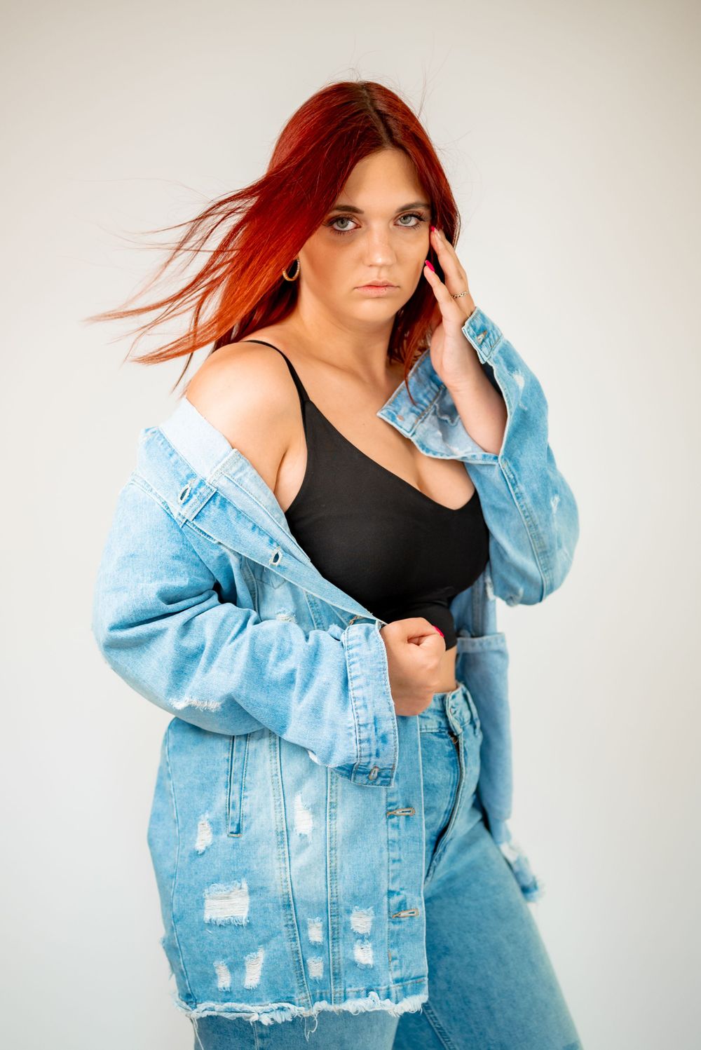Fashion model with flowing red hair wearing distressed denim outfit against white background.