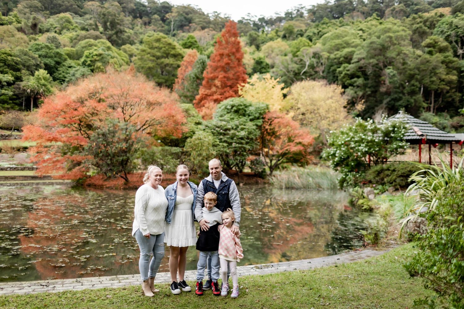 Family portrait in front of vibrant autumn trees and a peaceful lake in a serene park setting.