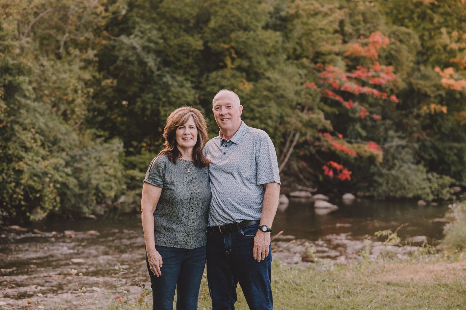 Couple poses together by a river with fall foliage creating a colorful natural backdrop.
