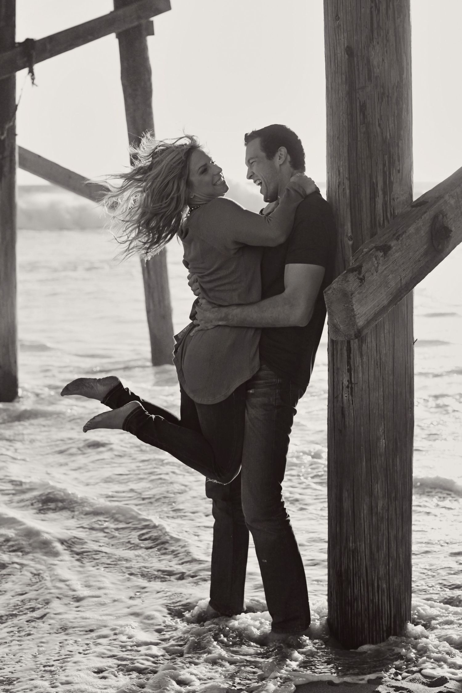 A playful couple poses against wooden pier posts at the beach in black and white.