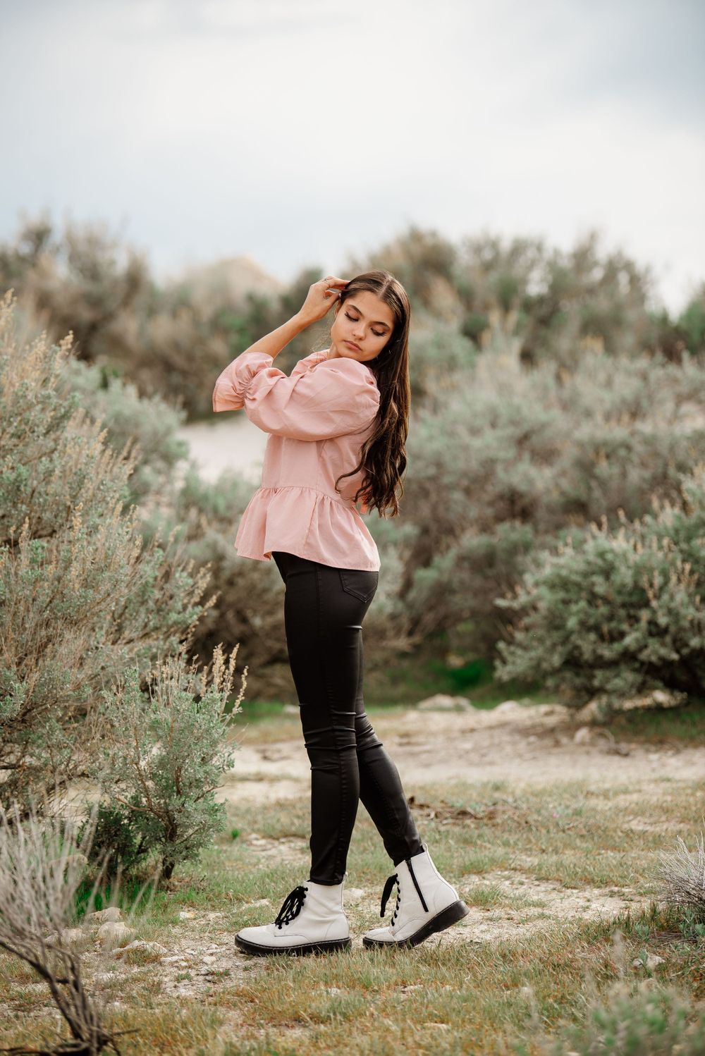 Person in pink top and black pants poses playfully in desert brush landscape.
