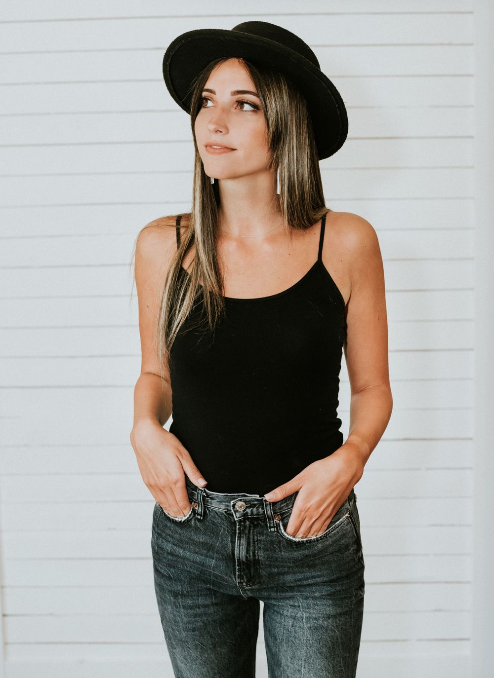 Casual fashion portrait featuring a black tank top, jeans and wide-brim hat against a white background.