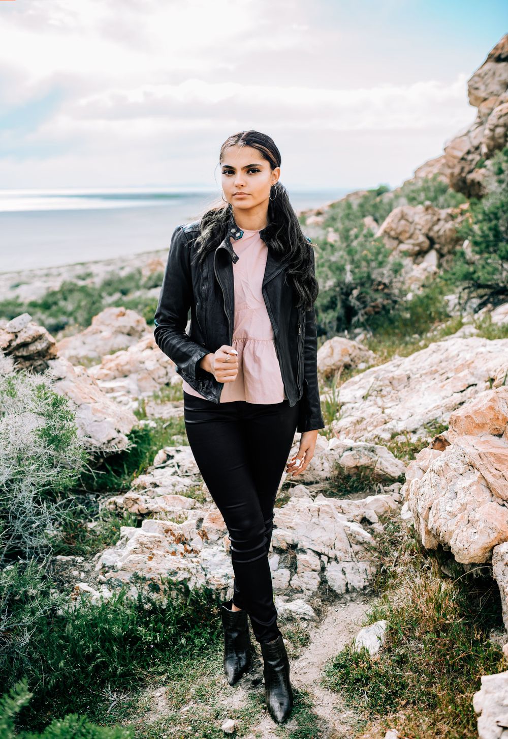 Person in leather jacket and pink top poses on coastal rocky cliff overlooking ocean.