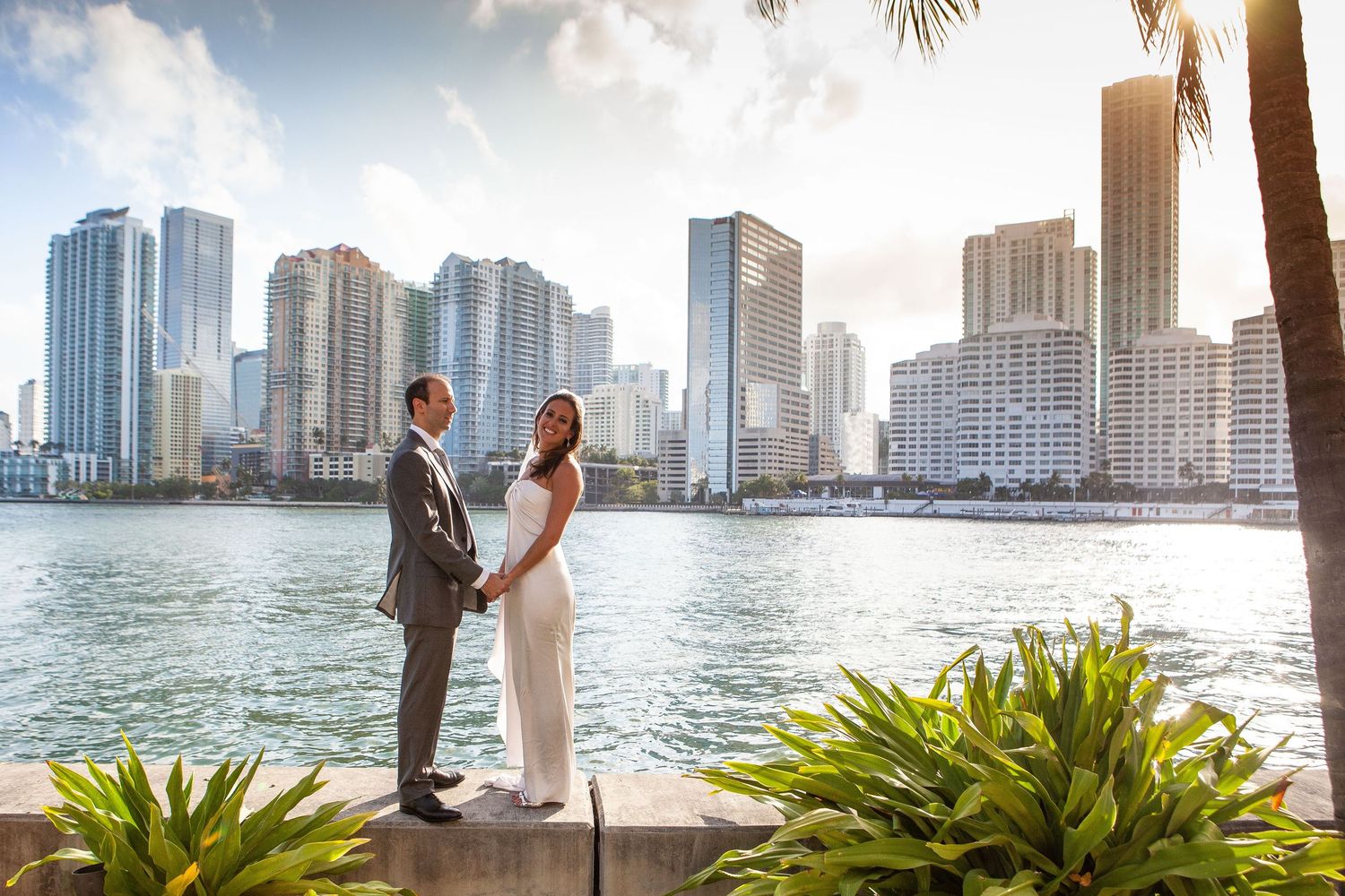 A couple stands by the waterfront in Miami with luxury high-rise buildings and palm trees in the background.