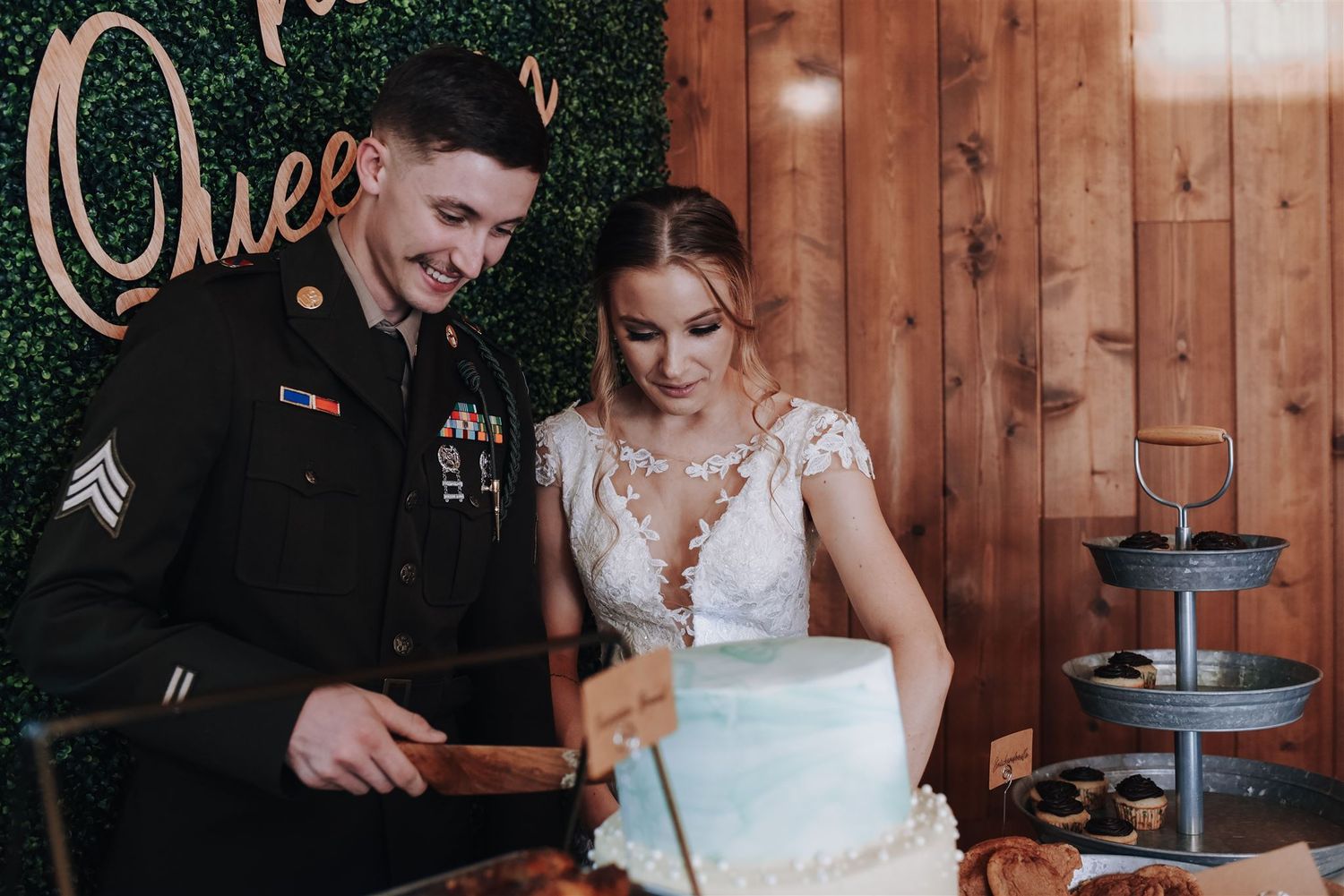 A couple cuts their wedding cake together on a dessert table with a decorative green backdrop.