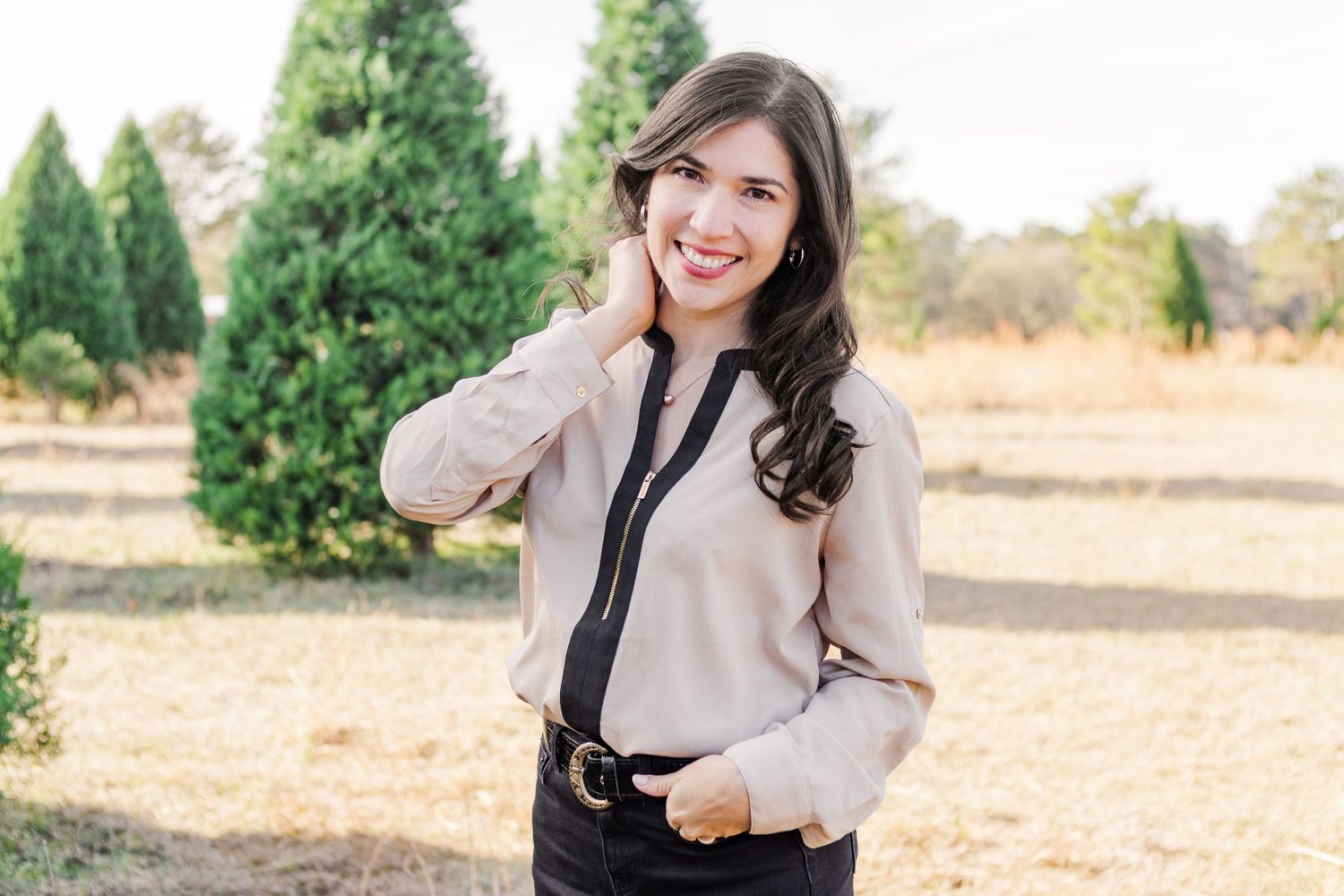 Woman in beige blouse and dark pants poses casually in outdoor setting with evergreens.