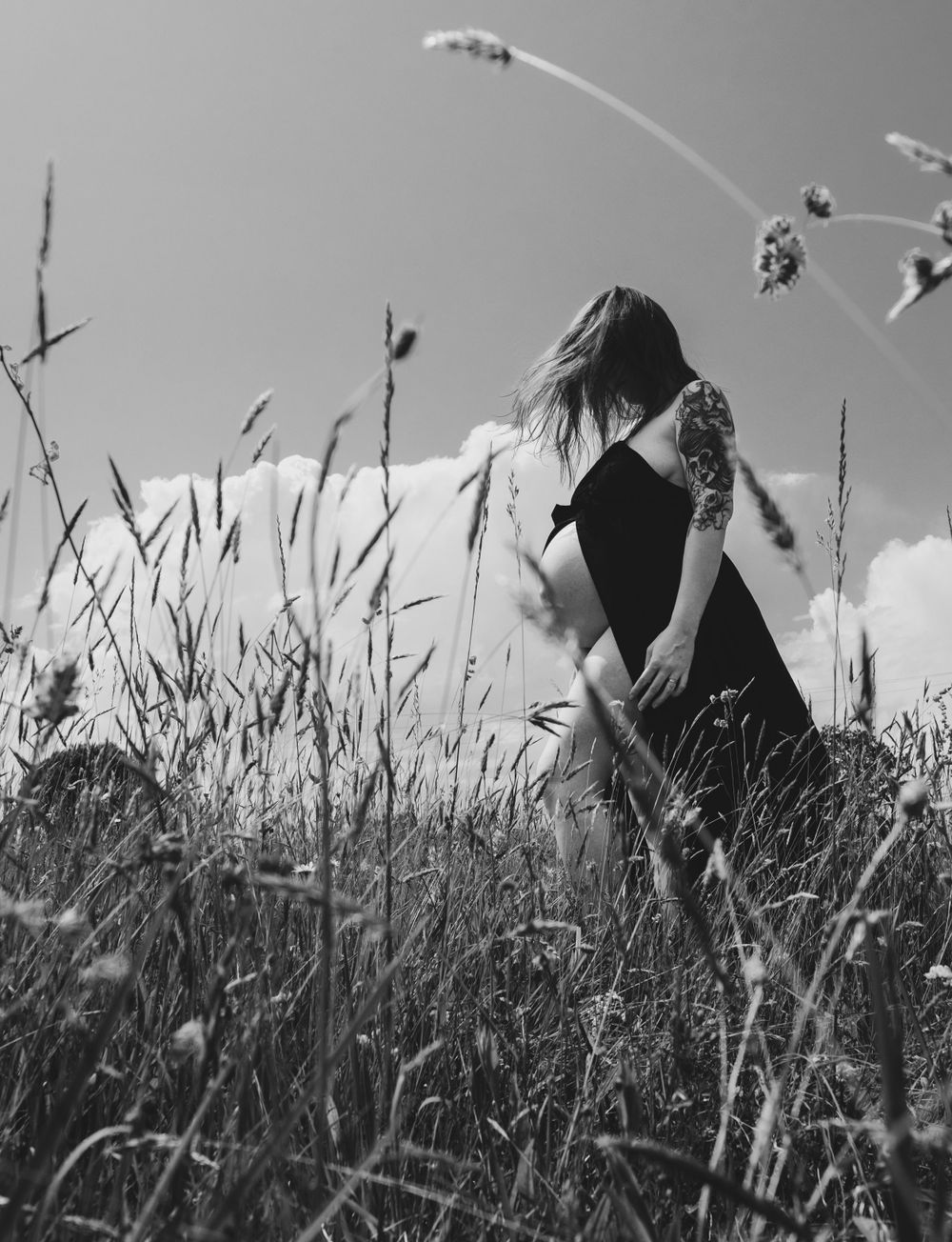 Black and white photo of person in dress standing among tall grass against cloudy sky.