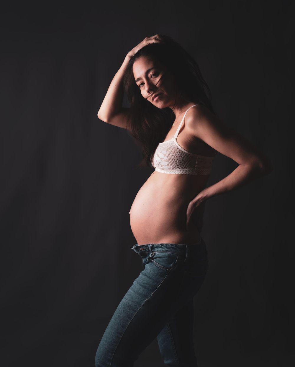 Artistic maternity portrait with expecting mother in jeans and crop top against dark background.