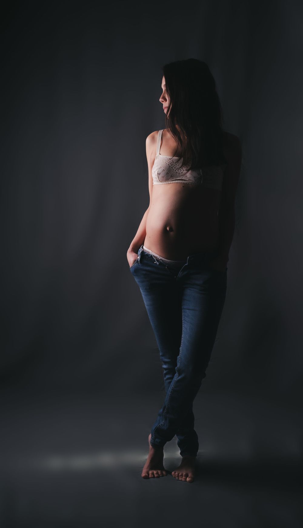 Moody studio portrait of woman in jeans and bralette posing against dark background.
