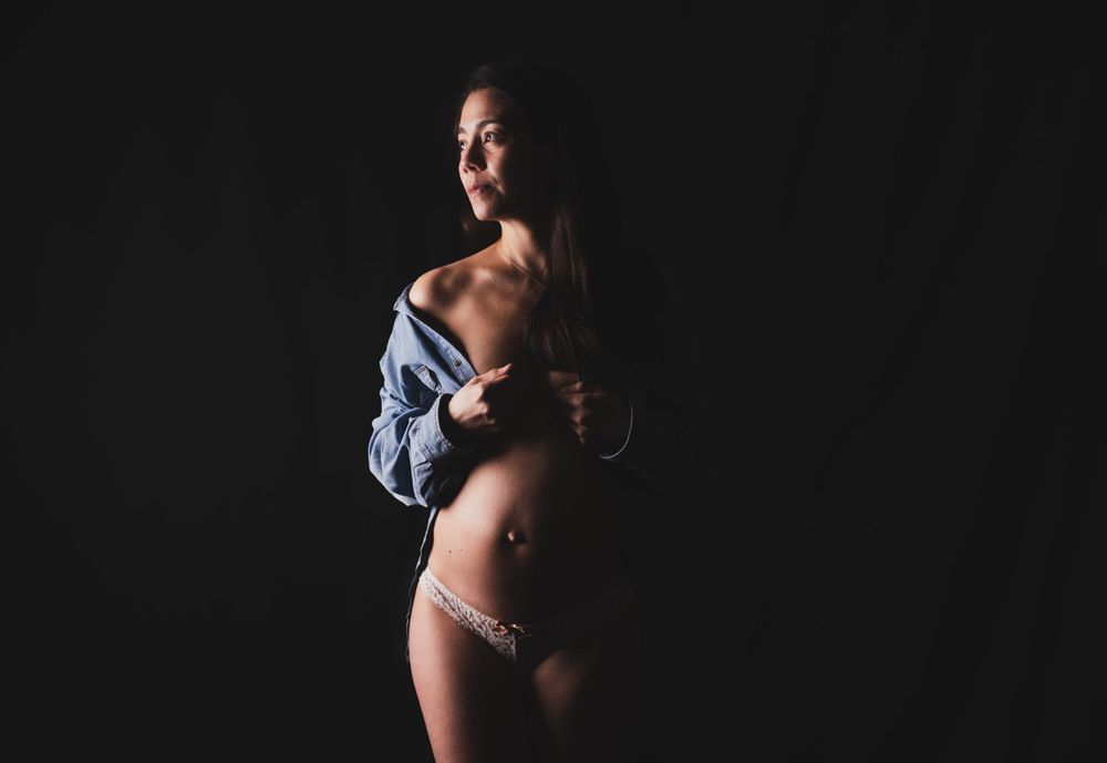 Intimate boudoir maternity photo with denim shirt against dark backdrop.