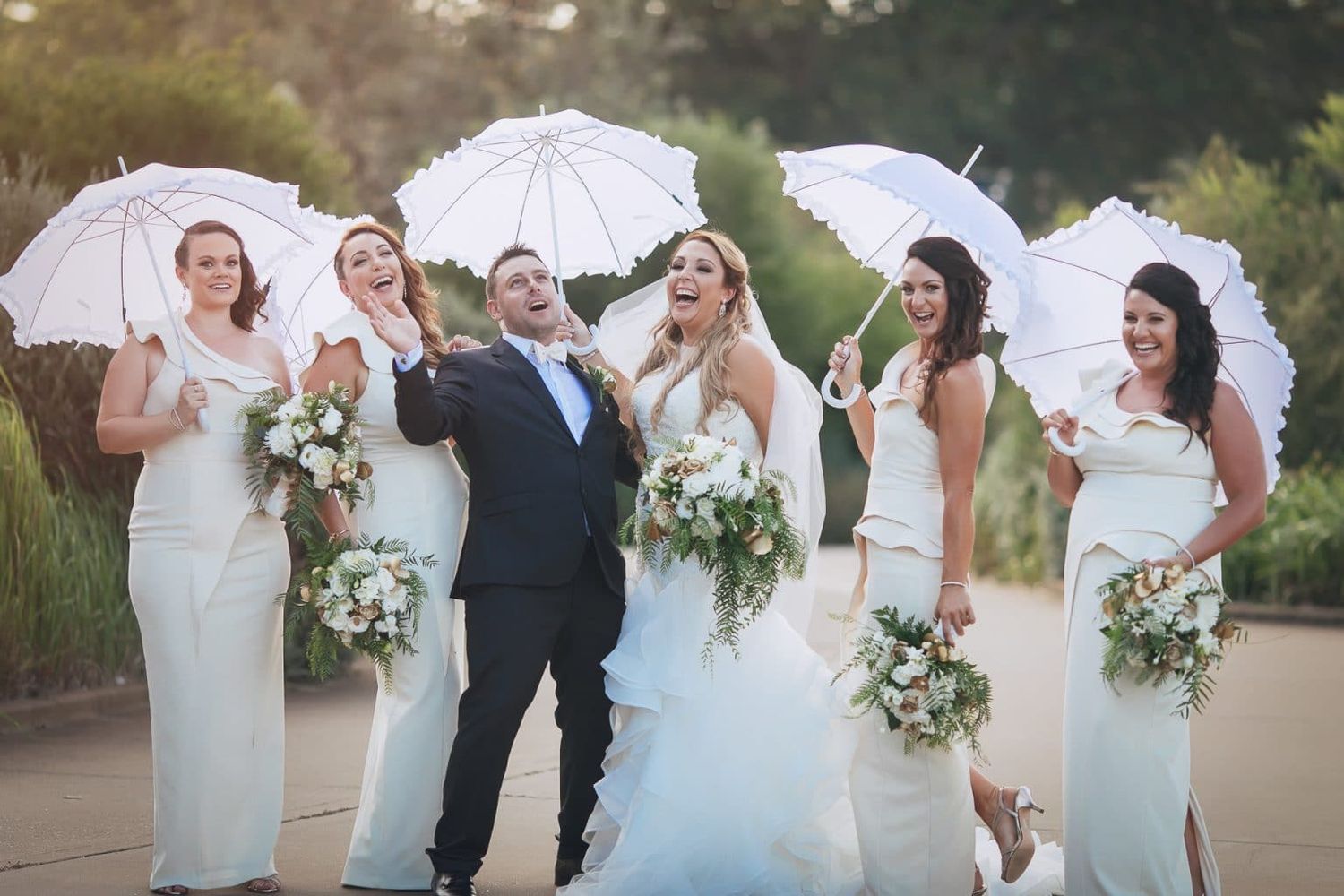 Wedding party celebrates outdoors with bridesmaids holding white parasols and floral bouquets beside smiling couple.