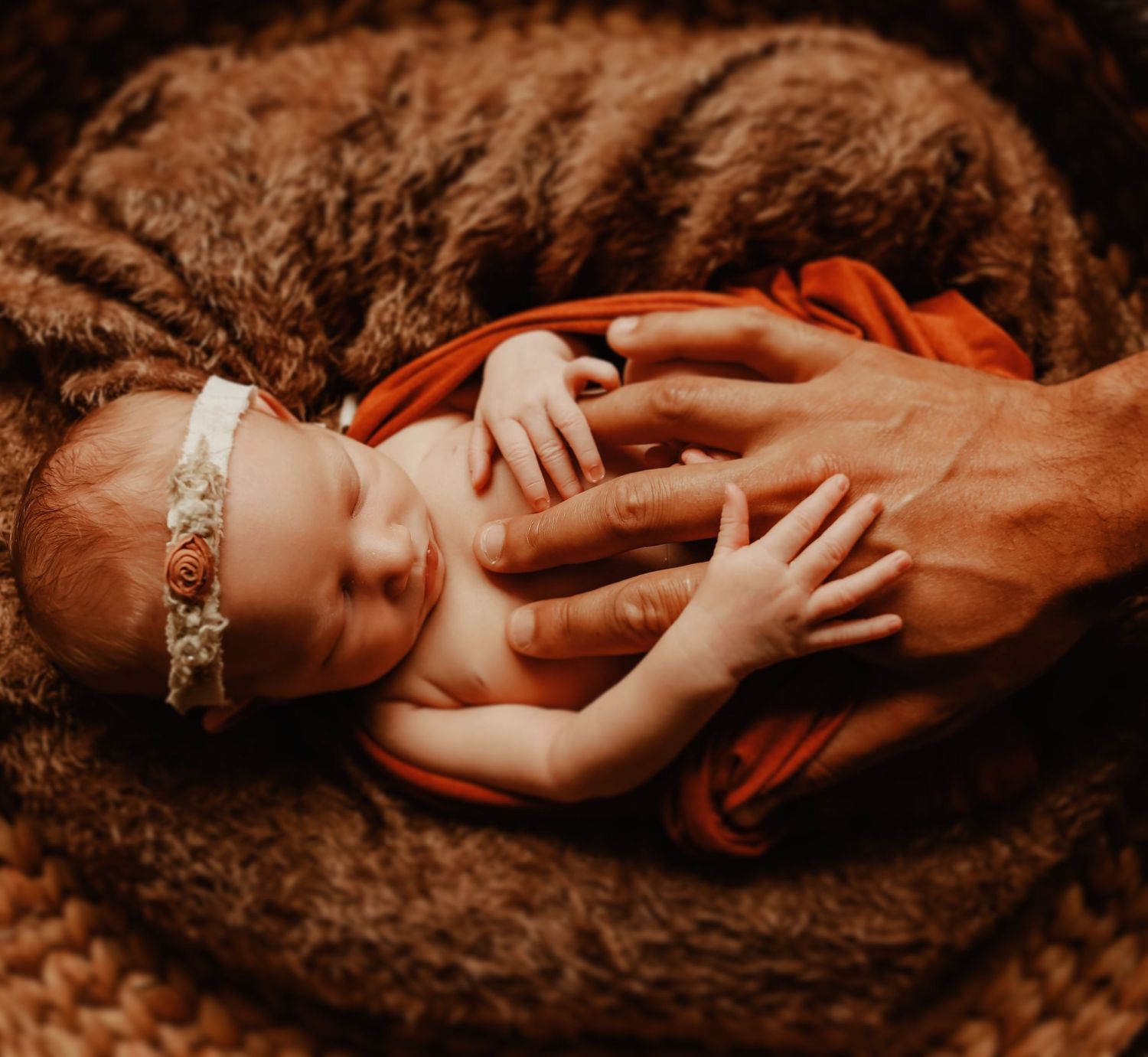 Newborn baby wrapped in orange fabric lies on brown textured blanket wearing delicate headband.