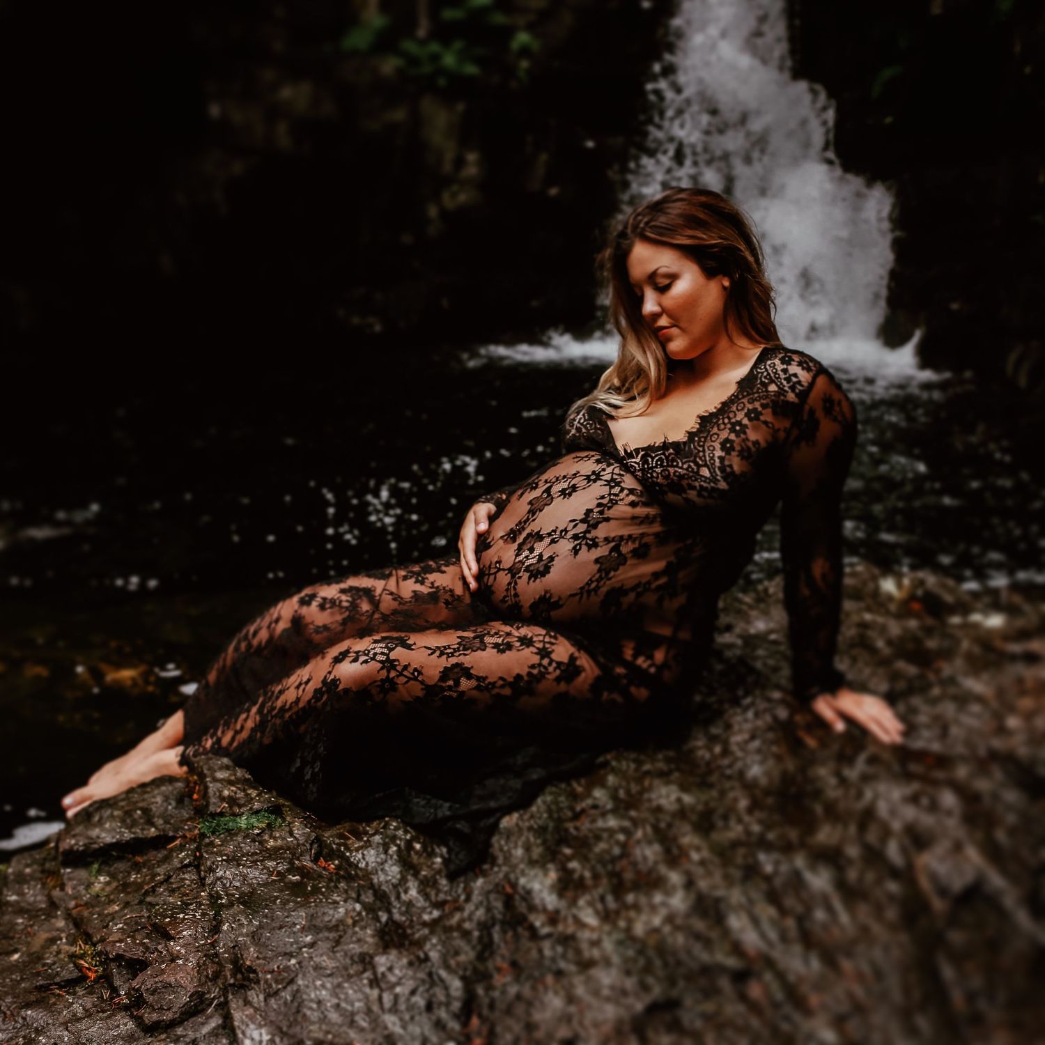 Woman in black lace dress poses gracefully on mossy rocks near a waterfall in moody forest setting.