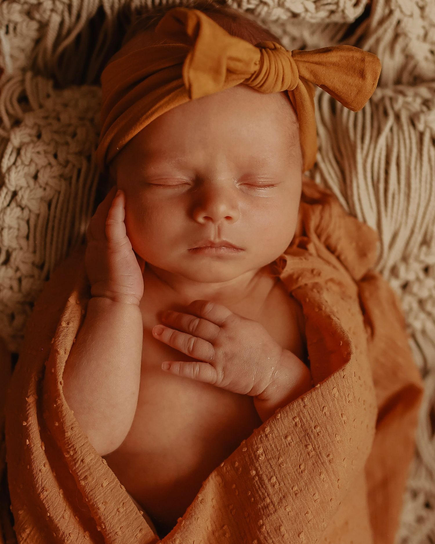 Sleeping newborn baby wrapped in peach-colored blanket with matching bow headband resting peacefully.