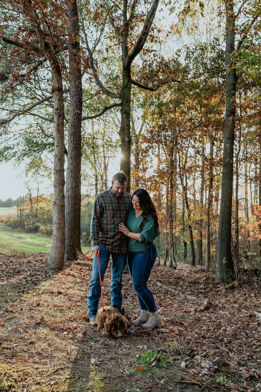 A couple takes autumn engagement photos in a wooded area with their dog.