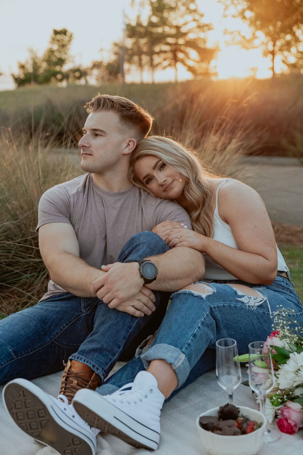A couple enjoys a sunset picnic with wine glasses and flowers on a blanket.