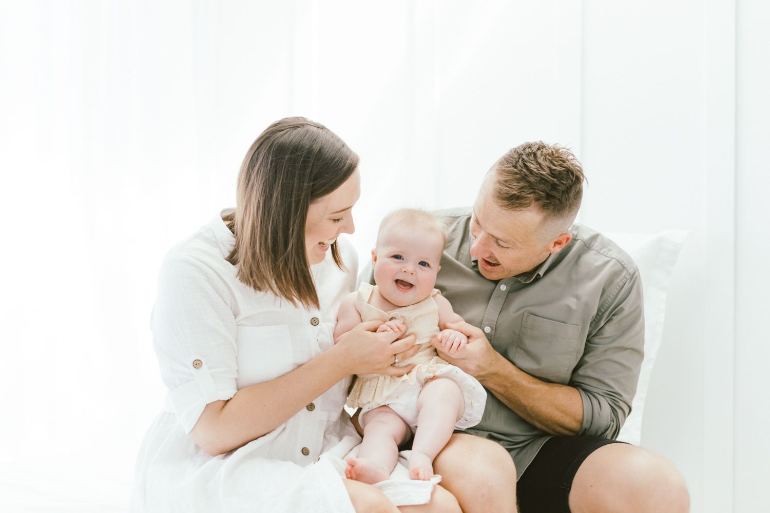Family having tender moment with baby wearing cream-colored outfit against white background.