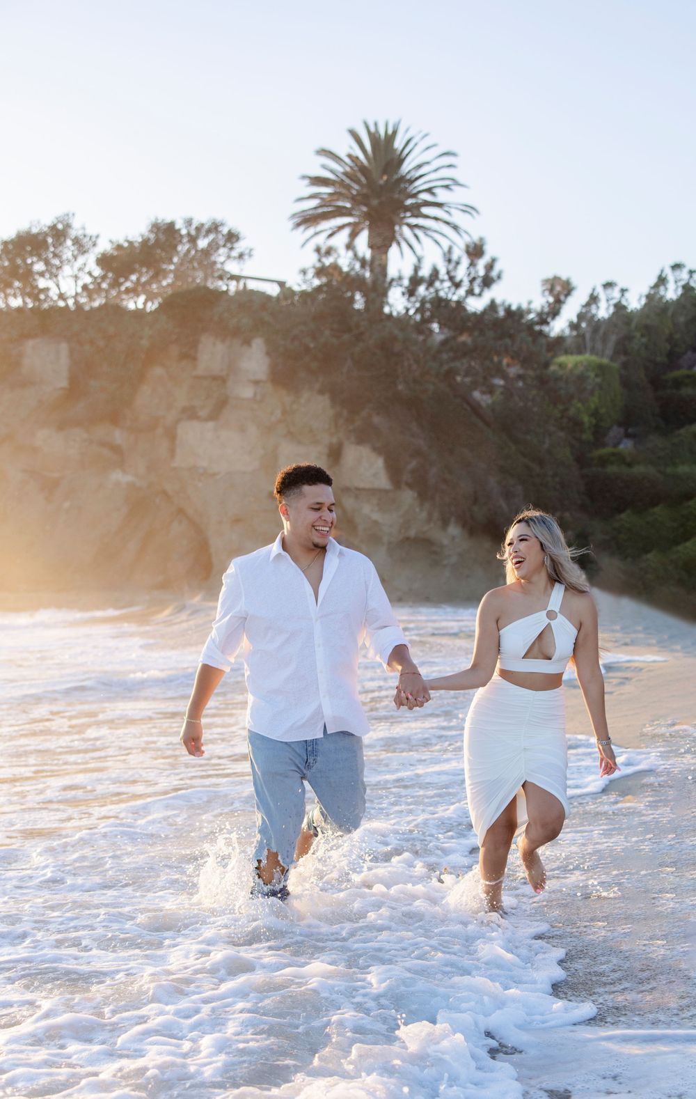 Couple splashing and playing in the ocean surf at sunset near a palm tree beach.