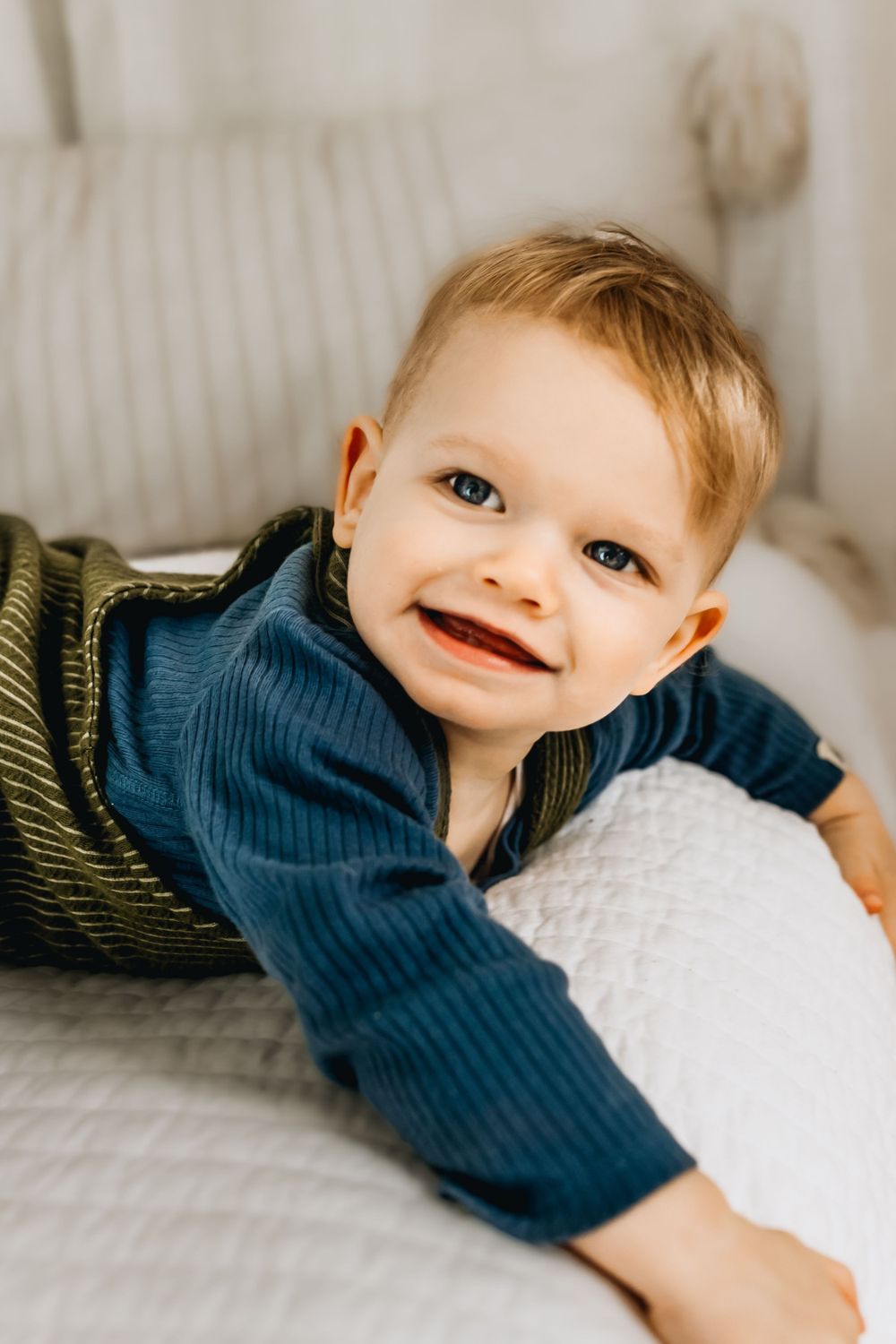 Smiling baby in blue outfit laying on white bedding.