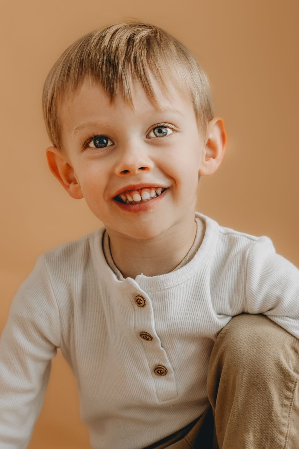 Child in white henley shirt smiling warmly against brown background.
