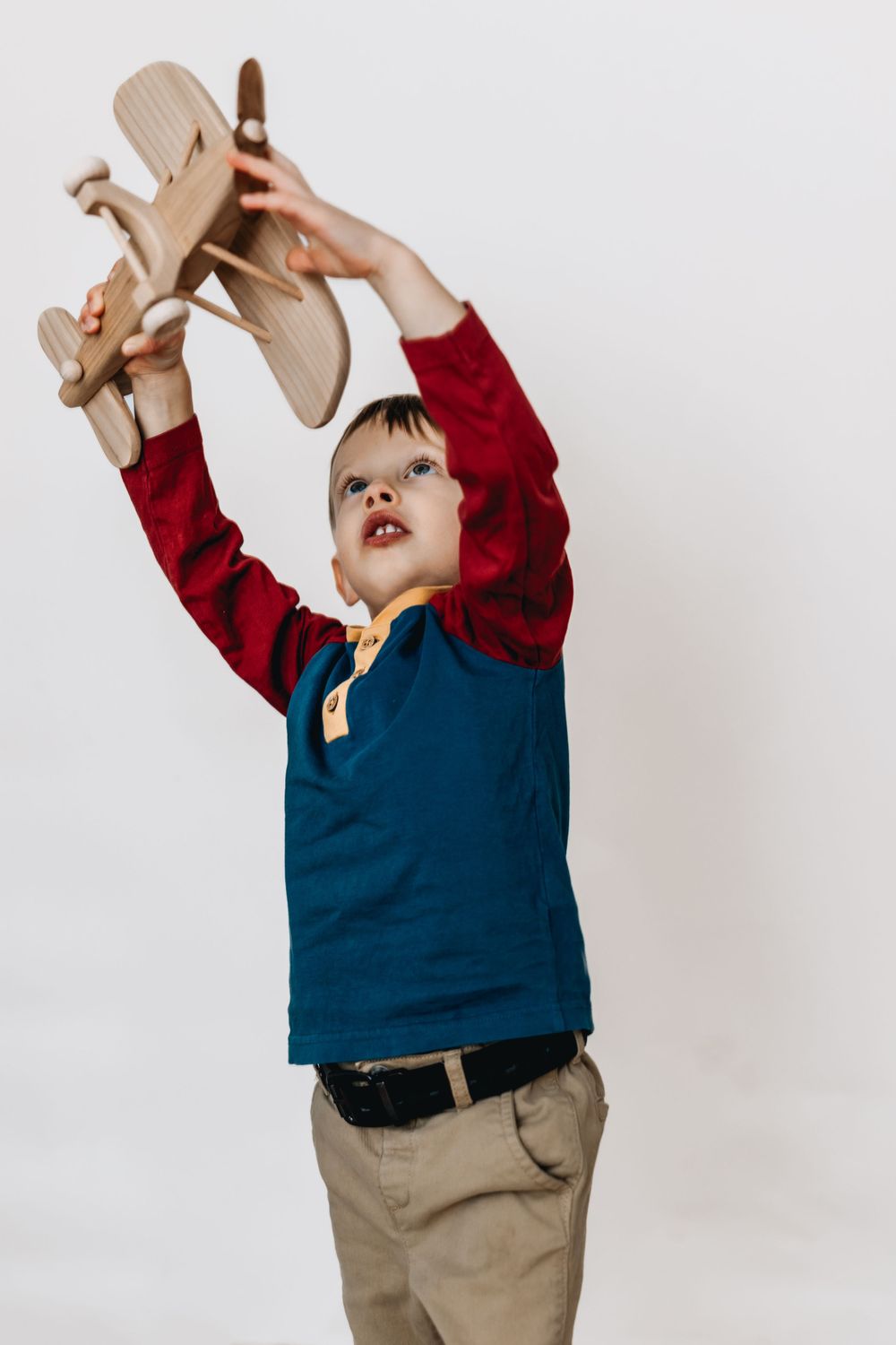 Child in blue and red top playing with wooden toy airplane against white background.