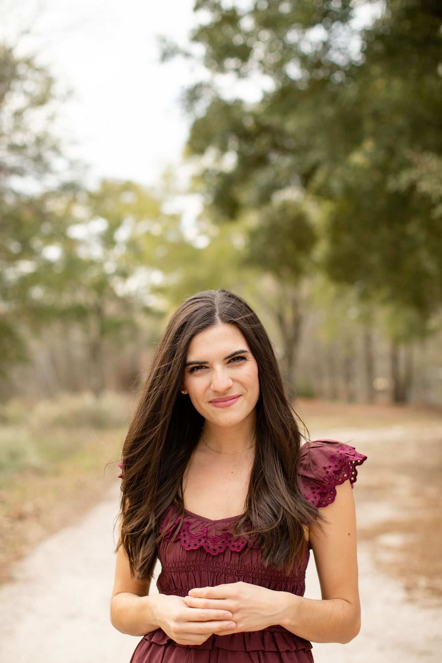 Woman in burgundy dress smiling while standing on a dirt path surrounded by trees.