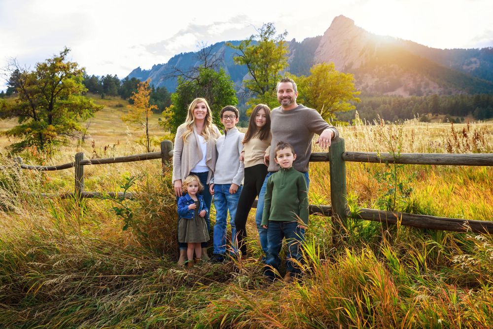 Family group poses along wooden fence with mountain backdrop during sunset.