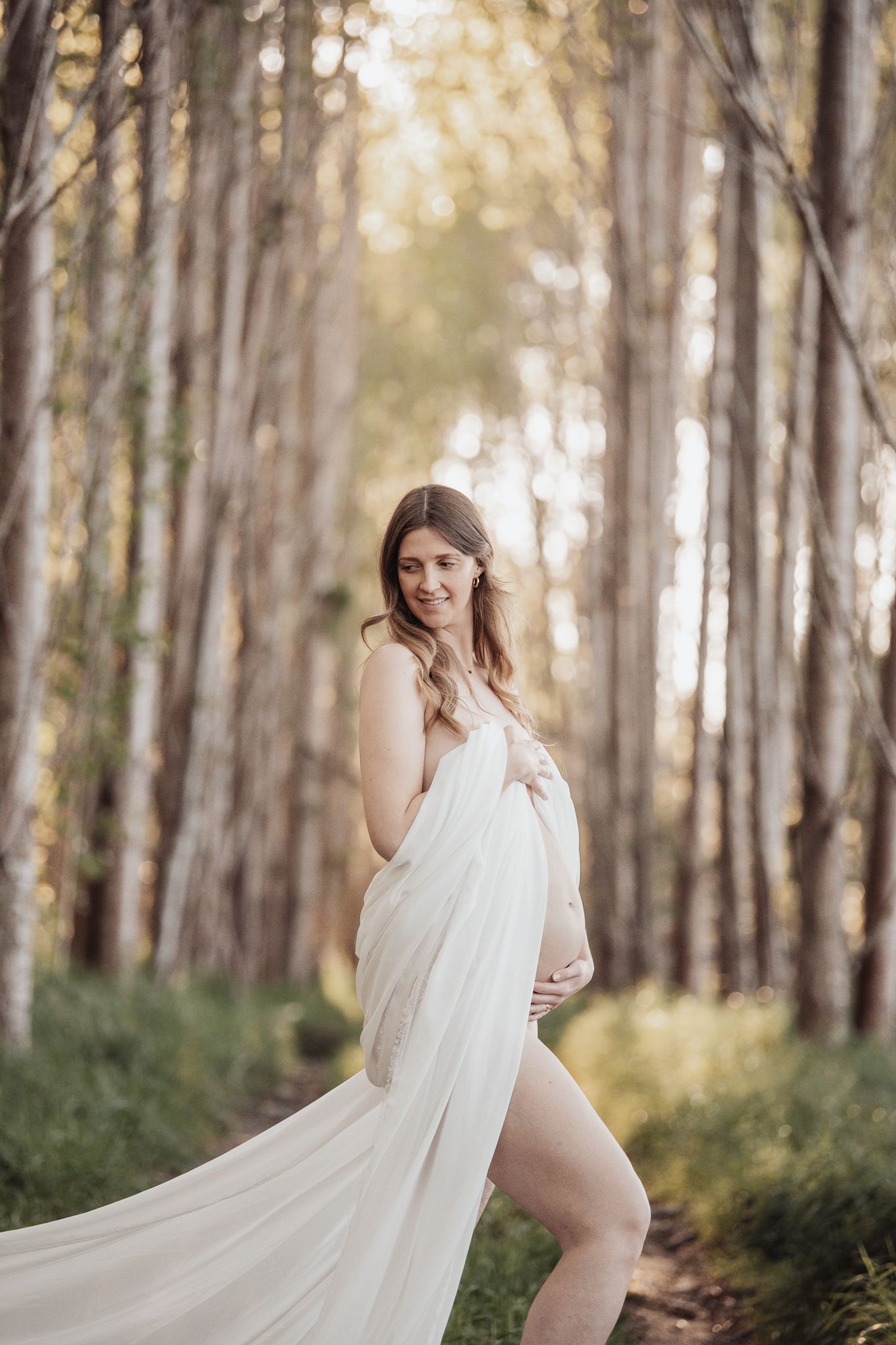 Ethereal maternity photo in forest with white flowing dress and golden sunlight through trees.