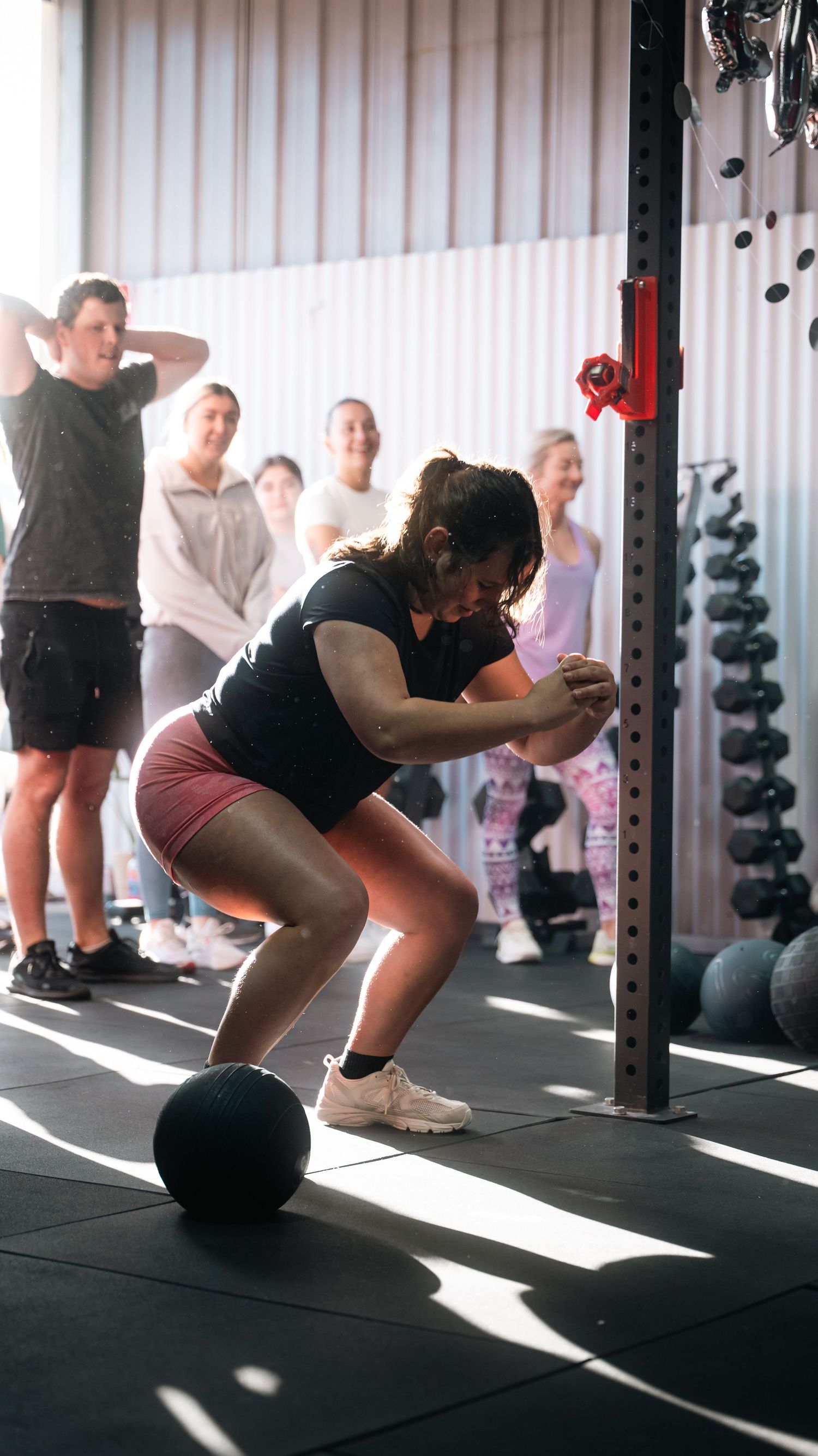 Athlete performs squats with medicine ball during group training session in sunlit gym.
