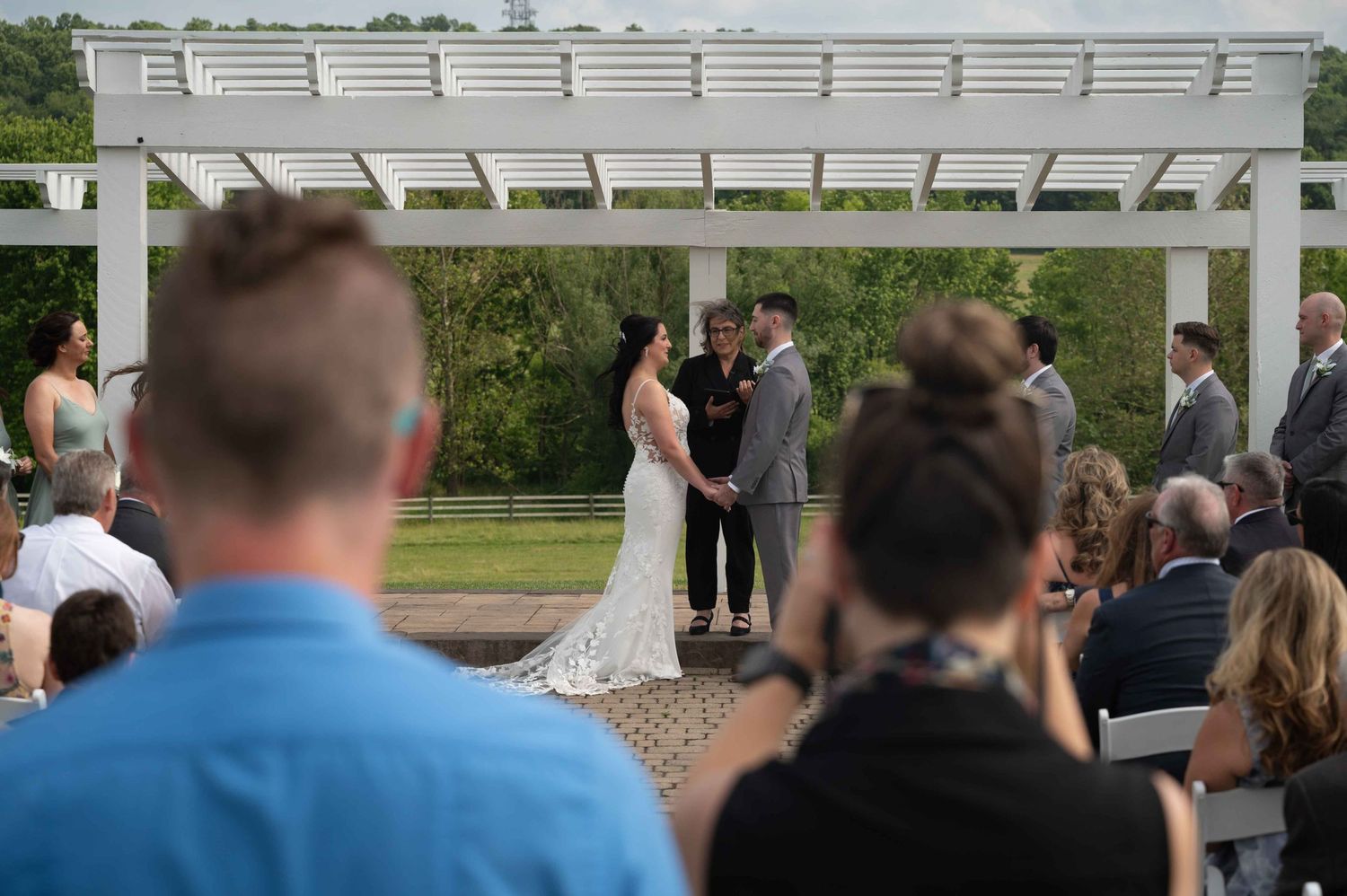 Wedding ceremony under white pergola with guests watching ceremony in outdoor setting.