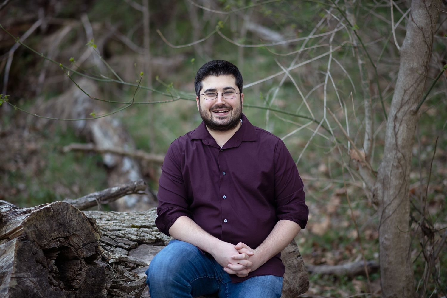 A person sits on a fallen log in a wooded setting wearing a purple button-down shirt.