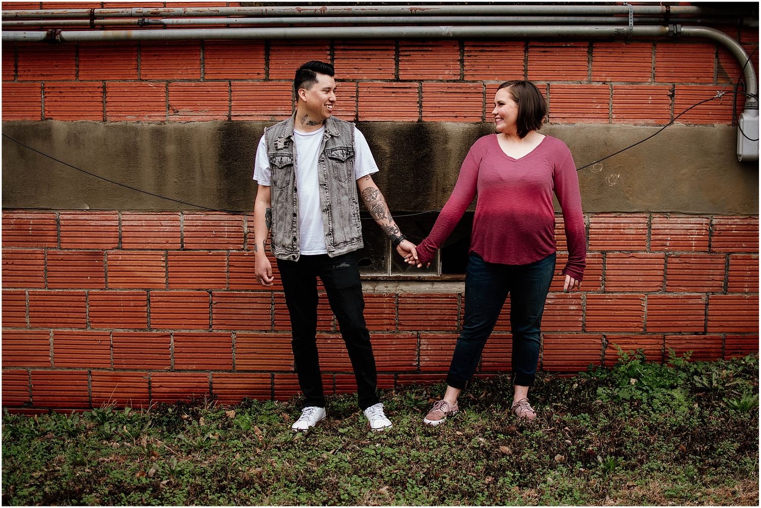 Couple holding hands and smiling against red brick wall in casual attire.