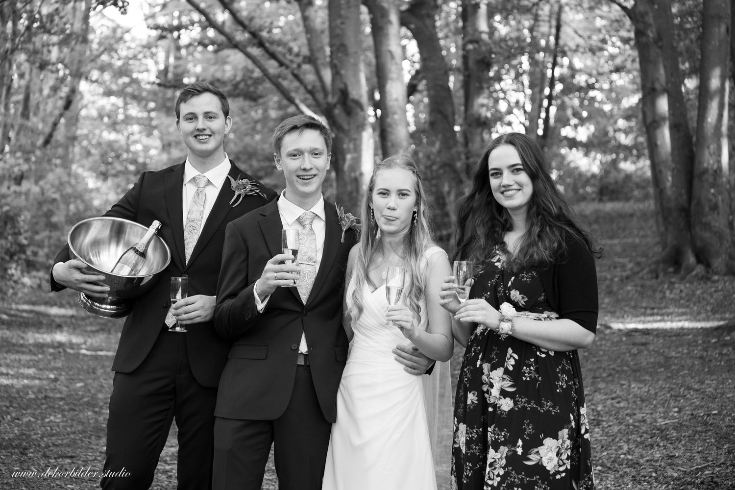 Black and white wedding photo of bride, groom, and two guests smiling outdoors holding champagne glasses.